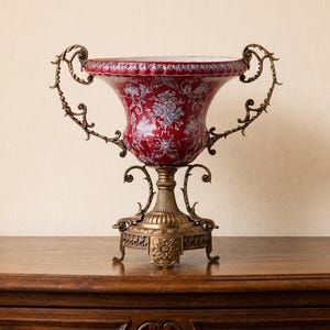 Peut inclure: Un vase décoratif rouge et or avec des poignées ornées et un motif floral. Le vase repose sur un socle doré détaillé, mettant en valeur un design antique. Le vase est exposé sur une surface en bois.