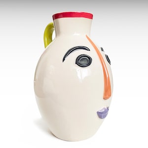 May include: A cream-coloured ceramic pitcher shaped like a face with a bright red rim, a lime green handle, a long orange nose, black arched eyebrows, black circular eyes, and purple lips. The pitcher has a smooth, glossy finish.