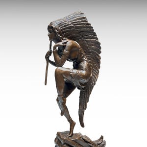 May include: Bronze sculpture of a Native American figure in a contemplative pose. The figure is seated, holding a staff, and wearing a detailed headdress. The sculpture is on a rocky base, with a neutral background.