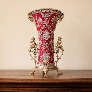 Può includere: Un vaso alto e svasato con un corpo rosso intenso e complessi motivi floreali argentati. Il recipiente è sostenuto da una base e una struttura in metallo dorato ornato, che include due piccole figure di cherubini sui lati.