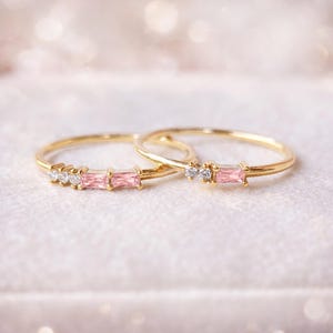 May include: Two elegant gold rings, each adorned with pink and clear gemstones. One ring showcases three pink baguette-cut stones and three clear round stones. The other ring features one pink baguette-cut stone and two clear round stones. Set against a soft, blurred backdrop.