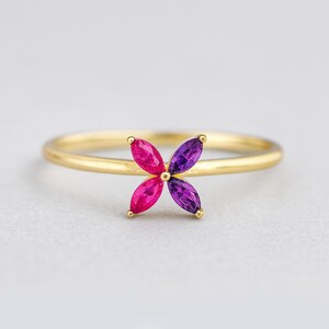 以下が含まれることがあります： ピンクと紫の宝石で作られた4枚の花びらのデザインが特徴のゴールドリング。リングは細いバンドで、明るい背景にセットされています。
