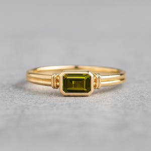 May include: A gold ring with a rectangular, olive-green gemstone. The ring has a double-band design and decorative elements on the sides of the gemstone. The ring is set against a gray background.