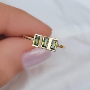 Könnte beinhalten: Ein goldener Ring mit drei rechteckigen, olivgrünen Edelsteinen, die horizontal angeordnet sind. Der Ring hat ein dünnes, goldfarbenes Band, und die Edelsteine sind in einem rechteckigen Rahmen gefasst.