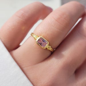 May include: A gold ring featuring a rectangular, pink gemstone. The ring has a delicate, detailed design with a thin band and decorative elements on the sides of the gemstone. The ring is a piece of jewelry.