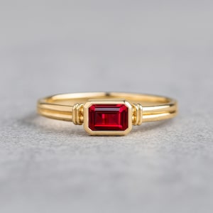 May include: A gold ring featuring a rectangular, deep red gemstone. The ring has a double band design and decorative elements on the sides of the gemstone. The gemstone is set in a bezel setting.