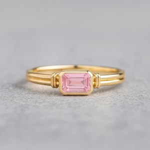 Könnte beinhalten: Ein goldener Ring mit einem rechteckigen, hellrosa Edelstein. Der Ring hat ein Doppelband-Design und dekorative Elemente an den Seiten des Edelsteins. Der Ring ist vor einem grauen Hintergrund platziert.