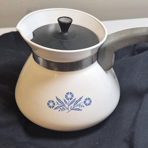 May include: A white teapot featuring a blue floral pattern on the front. The teapot has a black lid with a central knob, a metallic silver band around the upper section, and a gray handle extending from the side. The design is simple and functional.