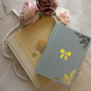 May include: Two journals with gold accents and decorative designs. The top journal is light blue with a gold bow and floral design. The bottom journal is cream with a gold floral design. The journals are surrounded by three pink roses.
