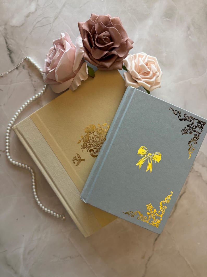 May include: Two journals with gold accents and decorative designs. The top journal is light blue with a gold bow and floral design. The bottom journal is cream with a gold floral design. The journals are surrounded by three pink roses.