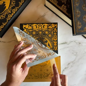 May include: A person is opening a decorative book with gold accents and a black cover. The book has a gold dragon illustration. Other books with gold embellishments are in the background. The book is on a white marble surface.