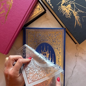 May include: A collection of hardcover books with gold foil designs. One book is blue with a castle design, another is magenta with a winged figure, and a third is black with an archer. A clear sticker is being peeled from the blue book.