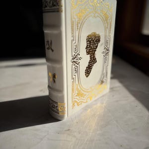 May include: White ceramic vase shaped like a book with gold accents and a silhouette of a woman's head on the front.