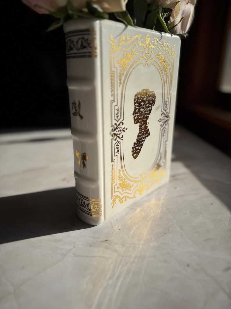 May include: White ceramic vase shaped like a book with gold accents and a silhouette of a woman's head on the front.