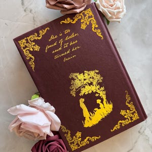May include: A burgundy hardcover book with gold accents and a gold silhouette of two people under a tree. The text on the cover reads "She is too fond of books and it has turned her brain".