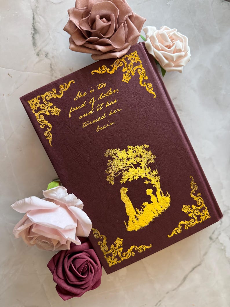 May include: A burgundy hardcover book with gold accents and a gold silhouette of two people under a tree. The text on the cover reads "She is too fond of books and it has turned her brain".