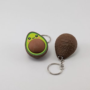 May include: Two avocado-shaped keychains. One keychain is a split avocado with a green pit and a smiling face. The other keychain is a whole brown avocado.