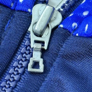 Może przedstawiać: Zbliżenie na srebrny suwak zamka błyskawicznego na tkaninie w niebiesko-białe wzory. Zamek błyskawiczny jest częściowo otwarty, odsłaniając zęby zamka.