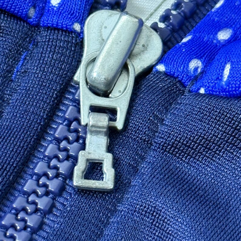 Może przedstawiać: Zbliżenie na srebrny suwak zamka błyskawicznego na tkaninie w niebiesko-białe wzory. Zamek błyskawiczny jest częściowo otwarty, odsłaniając zęby zamka.