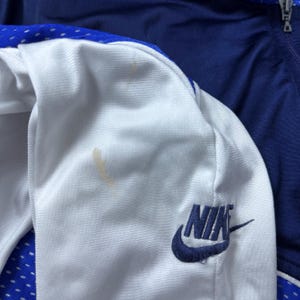 Może przedstawiać: Biała i niebieska kurtka sportowa z niebieskim logo Nike na rękawie.