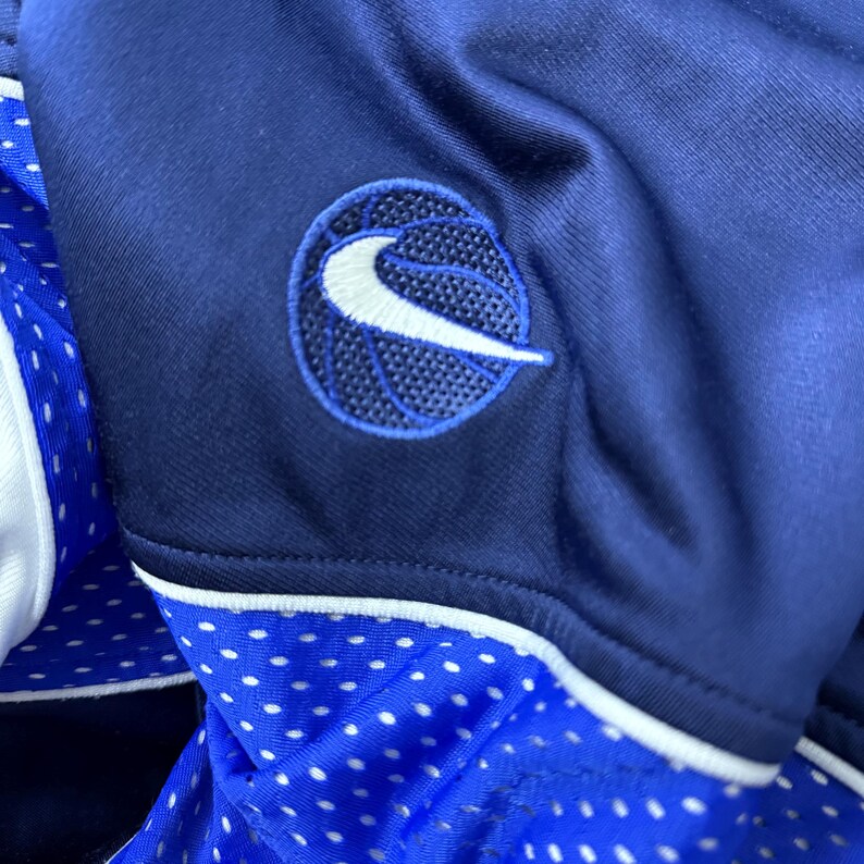 Może przedstawiać: Koszulka do koszyk&oacute;wki w kolorze granatowym z białym logo Nike i grafiką koszyk&oacute;wki z przodu. Koszulka ma biały pasek wok&oacute;ł szyi i rękaw&oacute;w. Koszulka jest wykonana z siateczki.