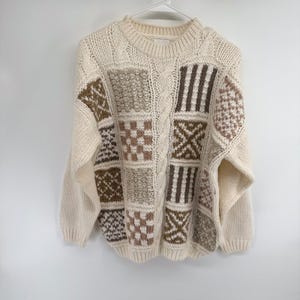 May include: Cream-coloured sweater with a crew neck and long sleeves. The front features a patchwork design with squares in shades of brown, tan, and cream. The sweater is hanging on a white hanger against a white background.
