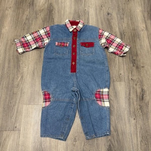 May include: A long-sleeve denim jumpsuit featuring a red, white, and black plaid pattern on the sleeves and knee patches. The garment includes a red button-down placket, a matching plaid collar, two chest pockets with red flaps, and metal snaps.