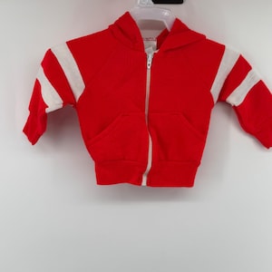May include: A bright red zip-up hoodie featuring a hood, long sleeves with wide white stripes, and two front pockets. The garment includes a white zipper closure running down the center and ribbed cuffs at the wrists and the bottom hemline.
