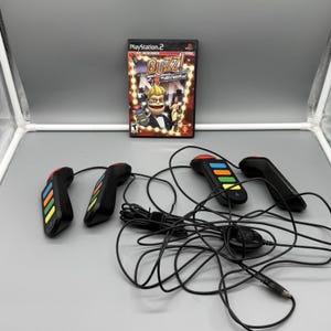 Pode incluir: Um jogo de PlayStation 2, "Buzz! Hollywood Quiz", com quatro controladores de jogo pretos. A caixa do jogo apresenta uma personagem de desenho animado com o título do jogo em estilo de tenda. Os controladores têm botões coloridos e tampas vermelhas.