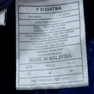 Może przedstawiać: Biała metka z czarnym tekstem, opisująca skład materiału i rozmiar odzieży. Metka zawiera napis "F 51204TWA", "RNS 86324", "CAM 05653", "AFCR NME 920220 KLA", "BODY: 100% POLYESTER", "OVERLAY: 100% NYLON", "TISSU D'EXTERIEUR: 100% POLYESTER", "APPLIQUES: 100% POLYAMIDE", "ONERPO: 100% POLYESTER", "PAROKG/S: 100% NYLON", "FABRIQUE EN MALAISIE", "HECHO EN MALASIA", "MADE IN MALAYSIA", "SIZE", "TAILLE", "TALLA", "M", "SEE REVERSE FOR CARE".