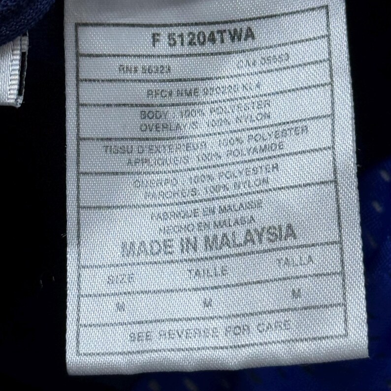 Może przedstawiać: Biała metka z czarnym tekstem, opisująca skład materiału i rozmiar odzieży. Metka zawiera napis "F 51204TWA", "RNS 86324", "CAM 05653", "AFCR NME 920220 KLA", "BODY: 100% POLYESTER", "OVERLAY: 100% NYLON", "TISSU D'EXTERIEUR: 100% POLYESTER", "APPLIQUES: 100% POLYAMIDE", "ONERPO: 100% POLYESTER", "PAROKG/S: 100% NYLON", "FABRIQUE EN MALAISIE", "HECHO EN MALASIA", "MADE IN MALAYSIA", "SIZE", "TAILLE", "TALLA", "M", "SEE REVERSE FOR CARE".