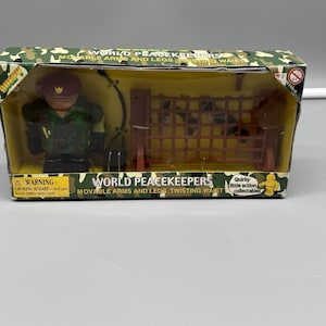 May include: A boxed "World Peacekeepers" action figure set. The figure wears a green uniform and a maroon beret. The set includes a brown net structure. The box has a camouflage pattern and text that reads "Movable Arms and Legs, Twisting Waist."