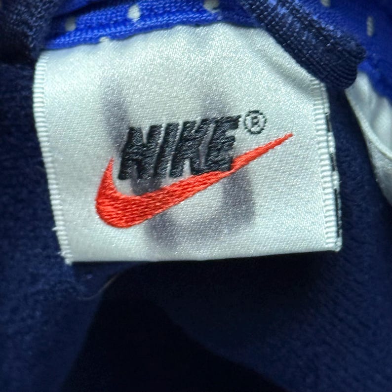 Może przedstawiać: Biała metka z napisem "NIKE" czarnymi literami i czerwonym logo swoosh. Metka jest przyszyta do granatowego materiału.