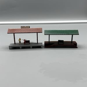 May include: Two miniature model train station platforms. The left platform has a brown roof, a sign reading PLASTICVILLE, and two small figures. The right platform features a green roof and a dark base. Both structures include a small bench.