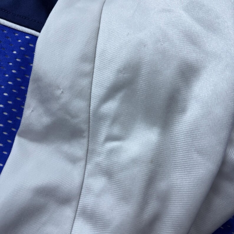 Może przedstawiać: Zbliżenie na białą koszulkę sportową z niebieskim i białym panelem z siatki. Tkanina ma subtelną fakturę.