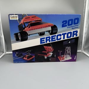 May include: A vintage Erector Set box featuring a red and black construction vehicle on a blue background. The box reads "200 Construction System" and "Erector". The Gabriel logo is visible. For ages 7 years and up.