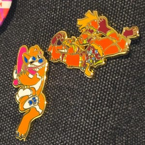 May include: Two enamel pins with gold trim. One pin features an orange cartoon character holding a pink object. The other pin depicts a similar orange character in a reclining pose. Both pins have blue, white, and red accents.