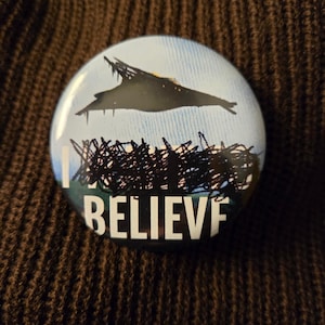 Könnte beinhalten: Ein runder Knopf mit blauem Hintergrund und einer schwarzen Silhouette einer fliegenden Untertasse. Der Text "I BELIEVE" ist in Weiß unter der Untertasse gedruckt.