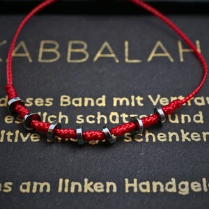 Op de afbeelding: Een rode koordarmband met zes kleine, donker metalen cilindrische kralen die gelijkmatig over het koord zijn verdeeld. De armband wordt getoond tegen een donkere achtergrond met gouden tekst, waaronder het woord KABBALAH in grote letters.