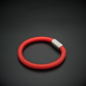 Könnte beinhalten: Ein rotes geflochtenes Armband mit einem silbernen Verschluss. Das Armband hat eine leuchtend rote Farbe und scheint aus einem dicht gewebten Stoff zu bestehen. Der Verschluss ist aus silberfarbenem Metall. Das Armband ist kreisförmig.