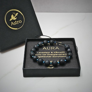 Könnte beinhalten: Ein Perlenarmband aus schwarzen, porösen Lavasteinperlen und kleinen metallisch blauen Akzentperlen. Das Armband liegt in einer schwarzen Schachtel mit dem Text AURA, Lavastein & Hämatit sowie weiterem deutschen Text auf der Einlegekarte.