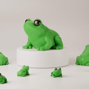 Op de afbeelding: Een groep van negen groene kikkerbeeldjes, met één grotere kikker die op een wit voetstuk zit. De grotere kikker kijkt omhoog, terwijl de kleinere kikkers in verschillende richtingen kijken. De tekst "TINY FROG" is zichtbaar bovenaan de afbeelding.