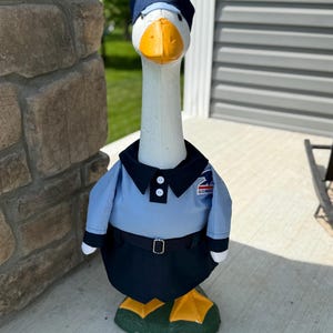 Könnte beinhalten: Eine skurrile Gänse-Statue in hellblauer Postuniform, komplett mit einer dunkelblauen Mütze mit amerikanischer Flagge. Die Gans hat einen weißen Körper, einen orangefarbenen Schnabel und grüne Füße. Die Statue steht auf einer Betonoberfläche.