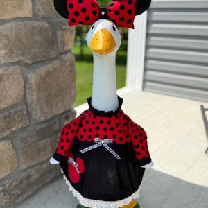 May include: A decorative goose statue wearing a red polka dot dress and a black bow with red polka dots. The goose has black ears and a yellow beak. The statue is painted with white, red, black, yellow, and green paint.