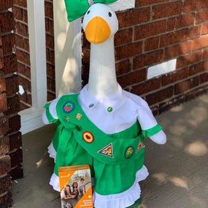 Puede incluir: Un ganso blanco vestido con un uniforme verde de Girl Scout con una faja blanca y insignias. El ganso lleva un lazo verde y está de pie sobre una superficie marrón. Una caja de galletas de mantequilla de cacahuete Girl Scout está en primer plano.