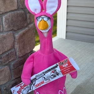 Può includere: Un'anatra di peluche con un costume da coniglietto rosa con occhiali, che tiene in mano una scatola di pallini Daisy 650. Il costume ha orecchie e piedi da coniglio. La scatola presenta un disegno di un fucile e la scritta "CARLINE".