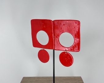 Puede incluir: Un par de pendientes rojos brillantes. Cada pendiente presenta una forma cuadrada con un gran recorte circular y un pequeño círculo rojo colgante. Los pendientes se exhiben en un soporte negro con una base decorativa.