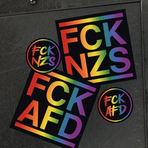 Könnte beinhalten: Drei regenbogenfarbene Aufkleber mit dem Text "FCK NZS" und "FCK AFD" in schwarzen Buchstaben. Die Aufkleber befinden sich auf einer dunkelgrauen Oberfläche.