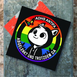 Könnte beinhalten: Schwarzer quadratischer Aufkleber mit einer cartoonartigen Panda-Figur mit Kapuzenpullover, vor einem Regenbogenhintergrund. Der Aufkleber trägt die weißen Schriftzüge "ADHS ANTIFA" und "ABGELENKT UND TROTZDEM DA". Ein Schmetterling ist ebenfalls abgebildet.