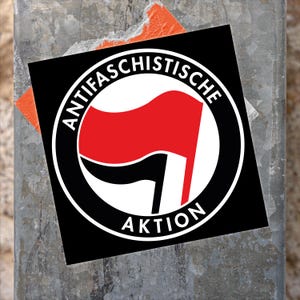 Könnte beinhalten: Ein schwarz-weißer Aufkleber mit dem Text "Antifa Schistische Aktion" in einem Kreis. Der Kreis enthält eine rot-schwarze Flagge mit weißem Hintergrund.